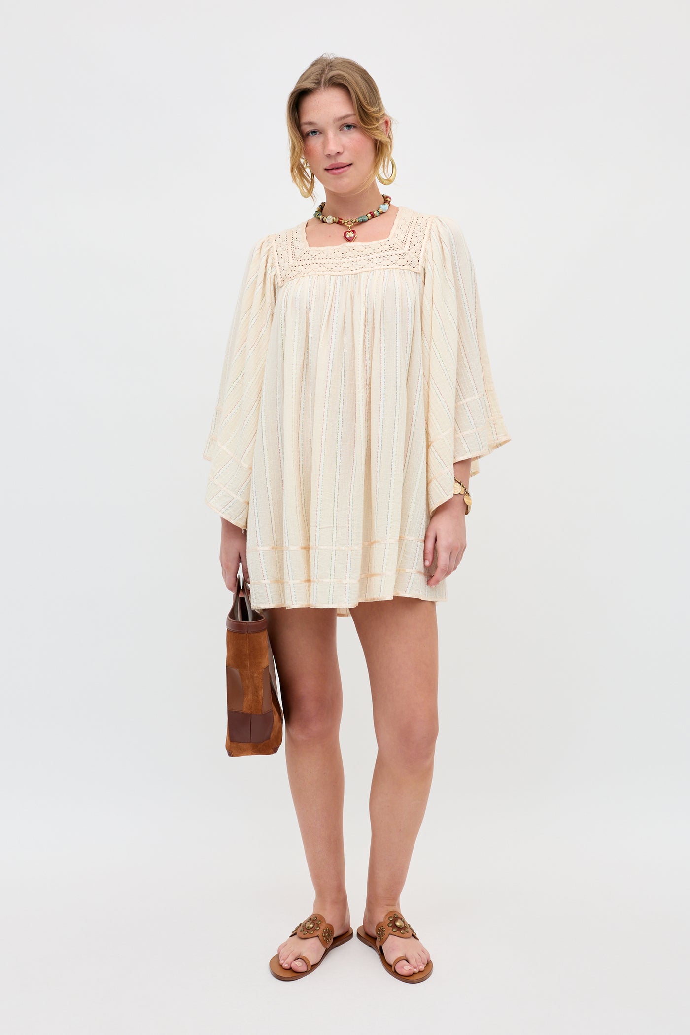 A woman stands against a white background wearing the Estele - Cream, a loose, long-sleeve dress, paired with brown sandals, a choker necklace, bracelets, and holding a brown handbag.