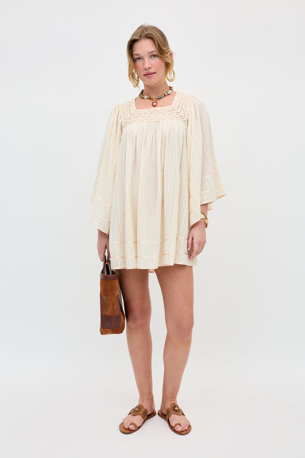 A woman stands against a white background wearing the Estele - Cream, a loose, long-sleeve dress, paired with brown sandals, a choker necklace, bracelets, and holding a brown handbag.