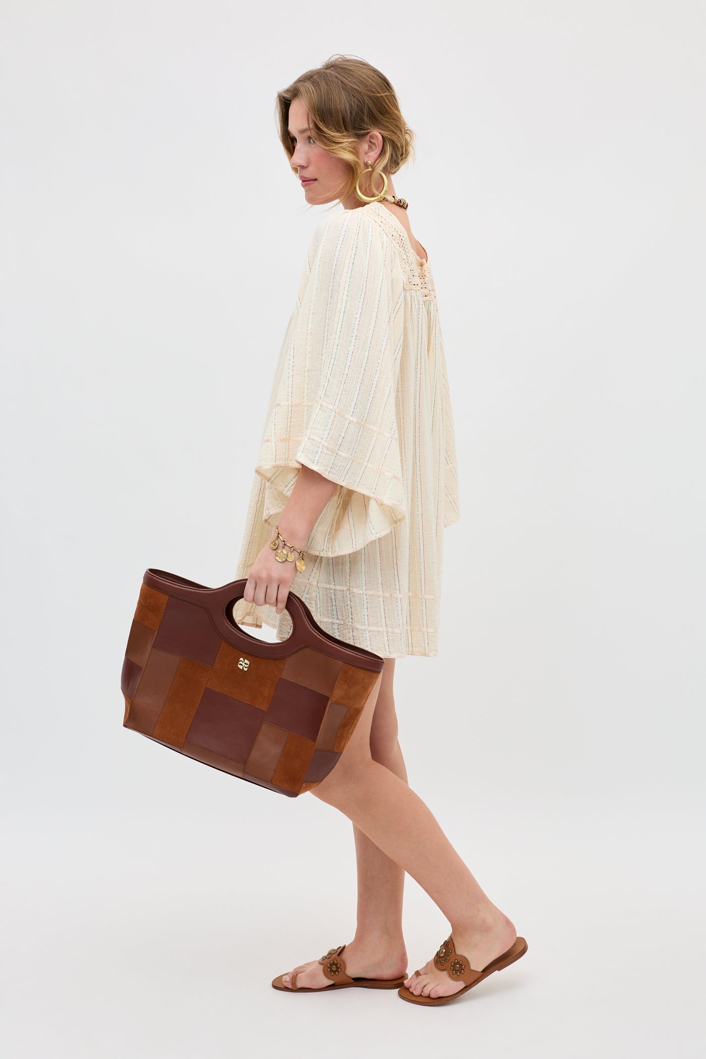 A woman stands sideways against a plain white background, wearing the Estele - Cream dress. She holds a large brown tote bag, pairs tan sandals and gold bracelets with her look, and has her hair loosely tied back.