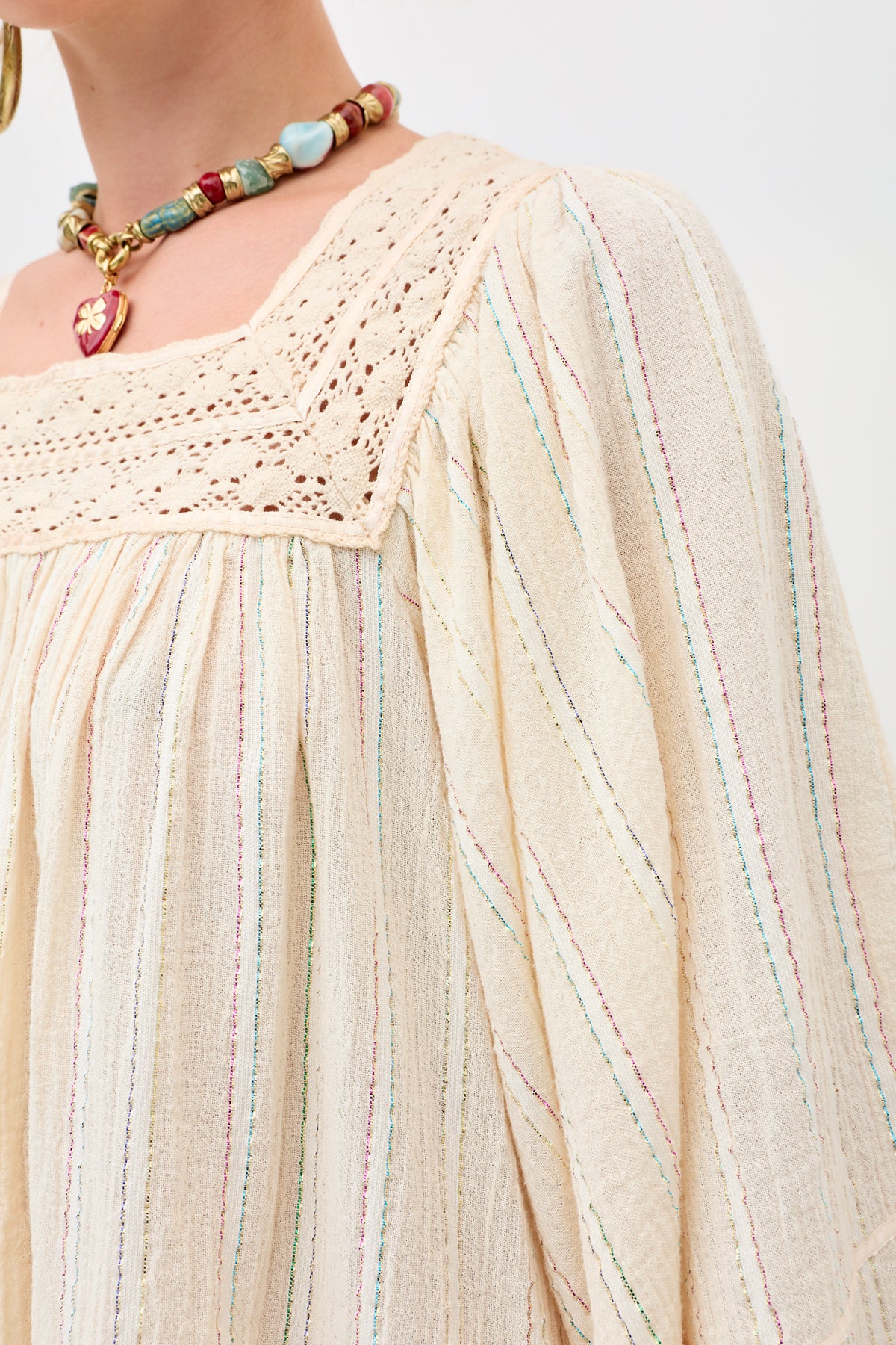 Close-up of a person wearing the Estele - Cream blouse, featuring multicolored vertical stripes and a square lace neckline, paired with a chunky beaded necklace. Only the shoulder, neck, and part of the face are visible.