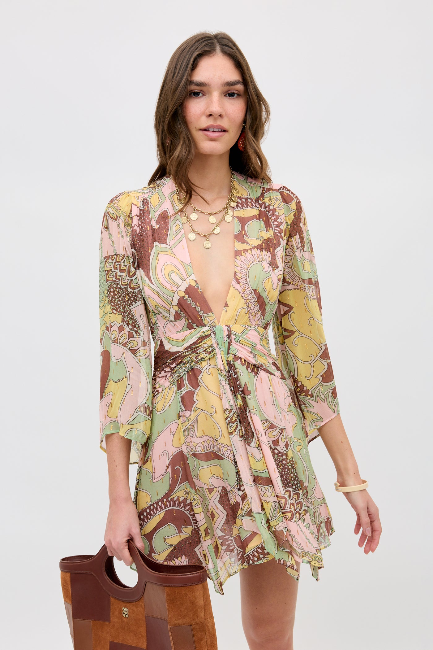 A woman wears the Clove - Bianca Abstract Green flowy V-neck mini dress with a colorful pink, green, and brown paisley pattern. She accessorizes with a brown patchwork handbag and layered gold necklaces.
