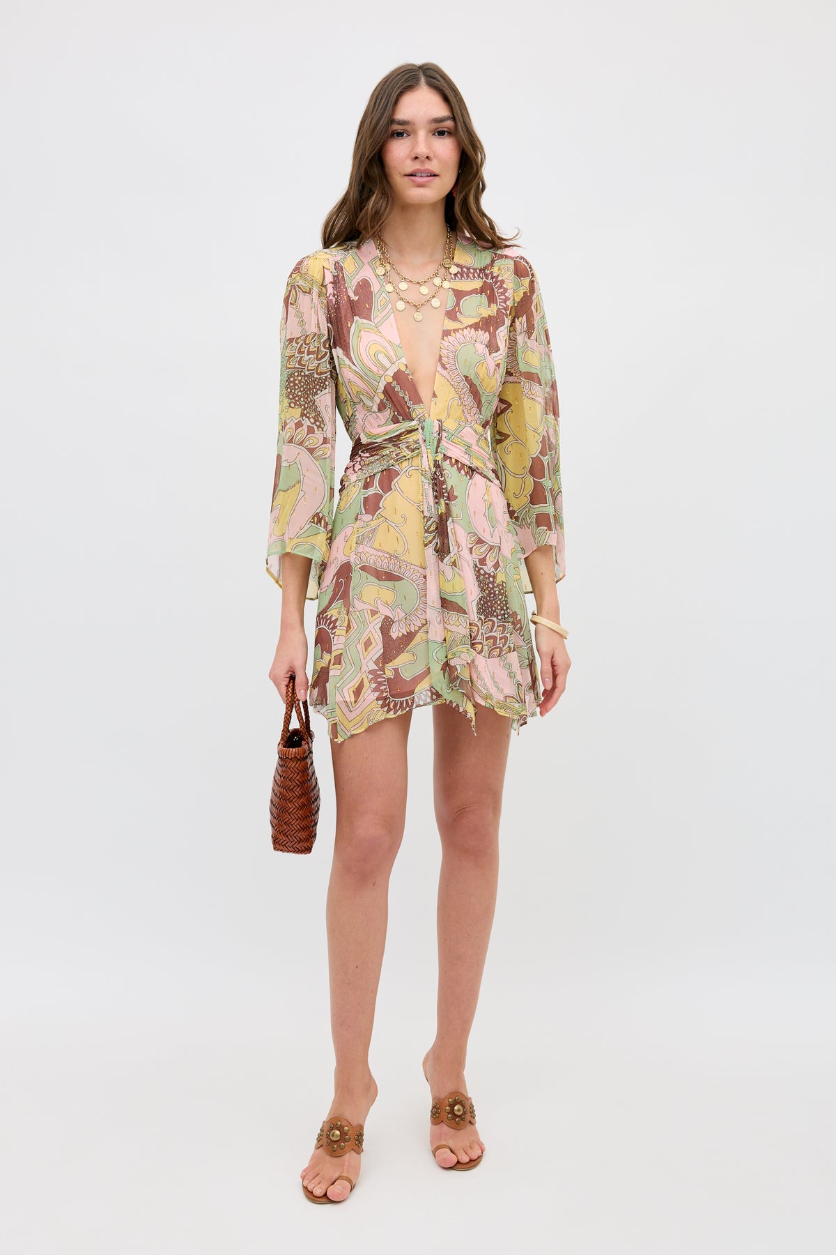 A woman stands against a plain background, wearing the Clove - Bianca Abstract Green, a flowy mini dress with a deep V-neck and three-quarter sleeves. She holds a small brown woven handbag and wears tan slide sandals.