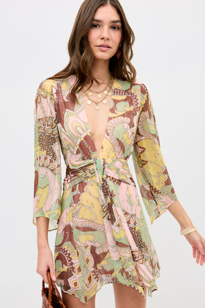 A woman wears the Clove - Bianca Abstract Green wrap dress with a deep V-neckline and flared sleeves, accessorized with layered necklaces, earrings, and bracelets, standing against a plain light background.