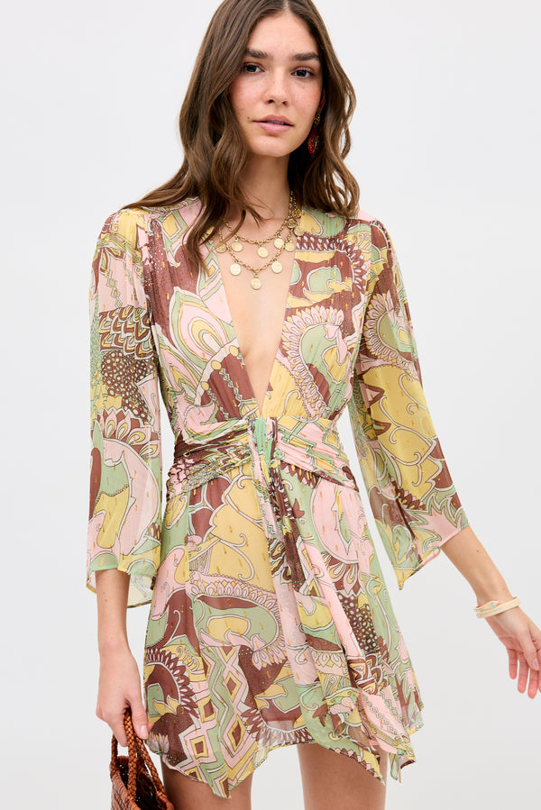 A woman wears the Clove - Bianca Abstract Green wrap dress with a deep V-neckline and flared sleeves, accessorized with layered necklaces, earrings, and bracelets, standing against a plain light background.