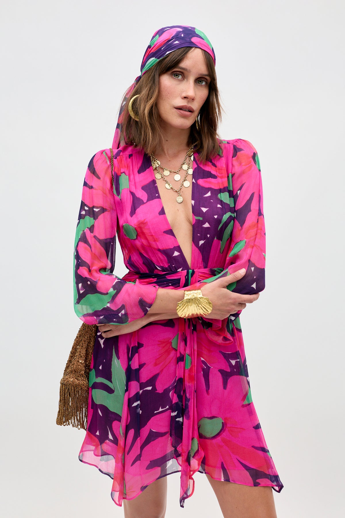 A woman poses with arms crossed wearing the Clove - Danish Floral Pink dress and a matching headscarf, accessorized with layered gold necklaces, a golden bracelet, and a brown fringe bag against a plain background.