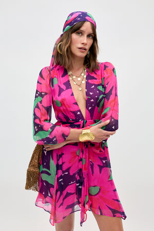 A woman poses with arms crossed wearing the Clove - Danish Floral Pink dress and a matching headscarf, accessorized with layered gold necklaces, a golden bracelet, and a brown fringe bag against a plain background.