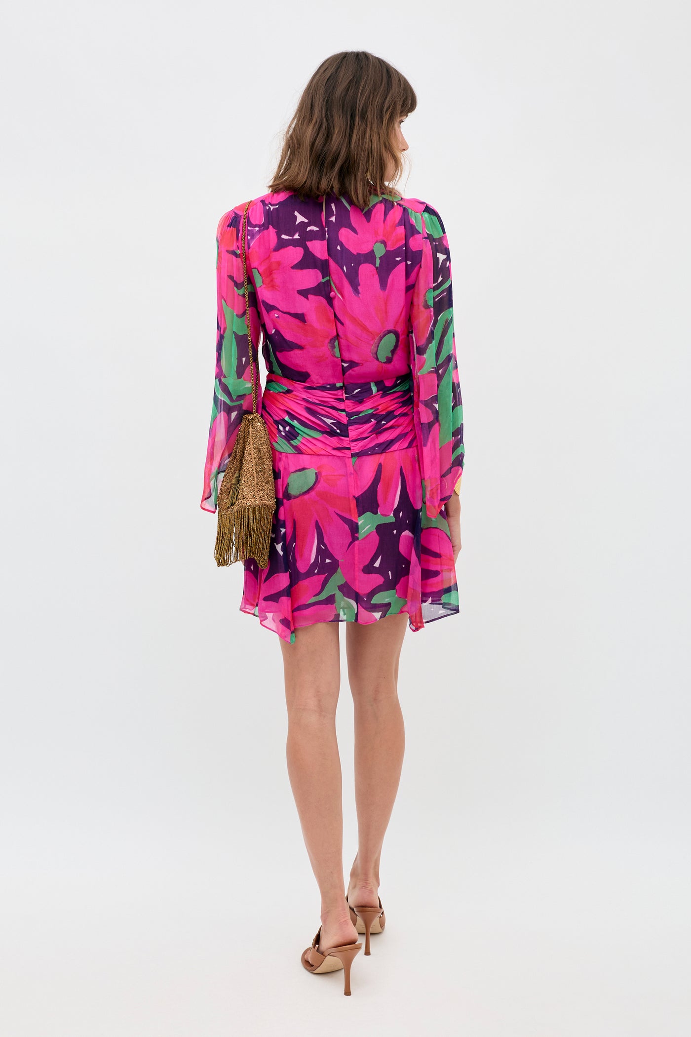 A woman faces away, wearing the Clove - Danish Floral Pink dress with a vibrant floral print. She carries a brown fringed bag and wears tan heeled sandals against a plain, light background.