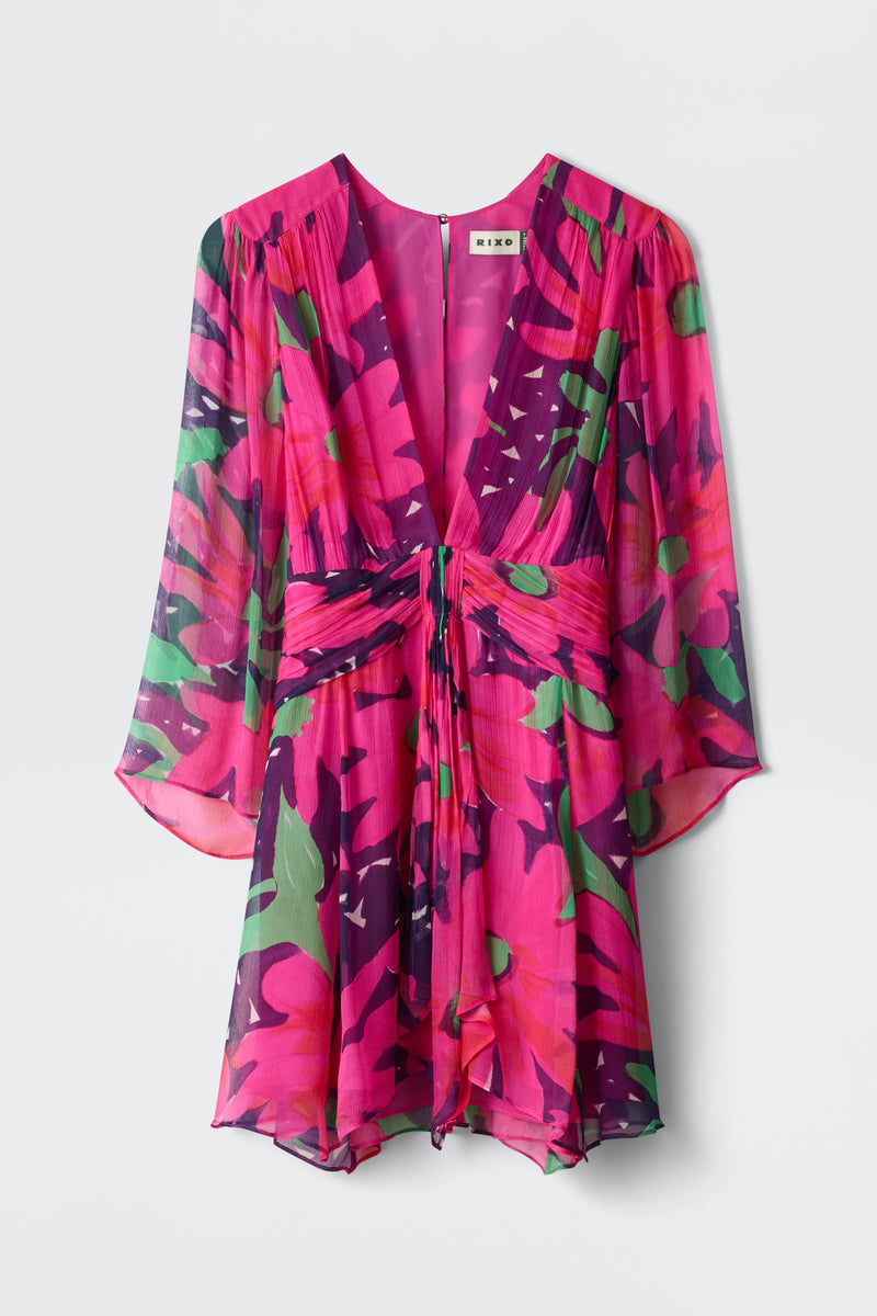 The Clove - Danish Floral Pink dress is a vibrant, sheer mini with a deep V-neck, waist tie, and flowing sleeves, featuring a bold pink, green, and purple floral print.