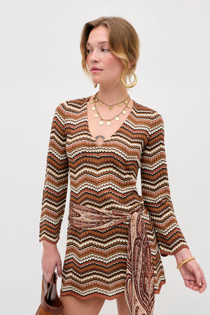 Wearing the Ivia - Maria Chevron Brown long-sleeve mini dress, layered gold necklaces, hoop earrings, and holding a small brown handbag, a person stands against a plain light background.