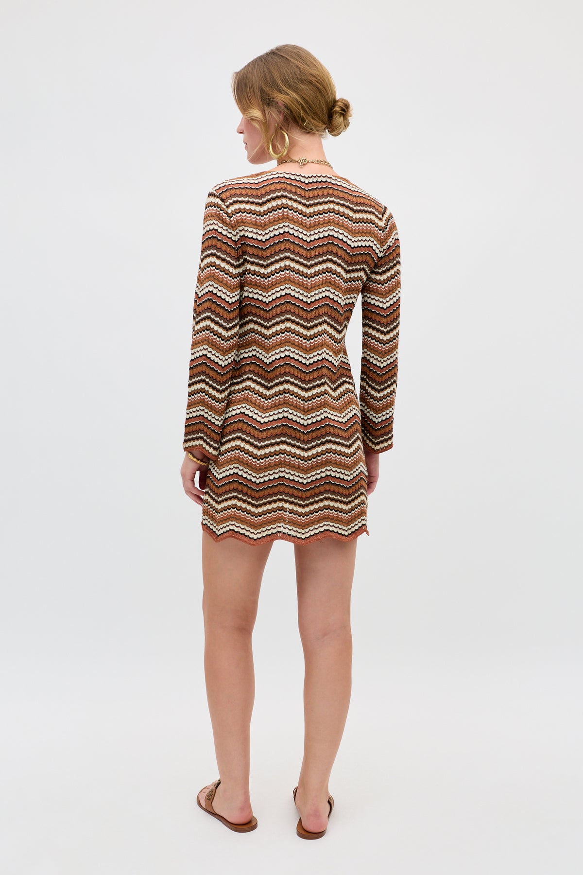 A woman with light brown hair in a low bun wears the Ivia - Maria Chevron Brown dress, a long-sleeve short dress featuring a brown, cream, and orange zigzag pattern. She pairs it with sandals and hoop earrings.