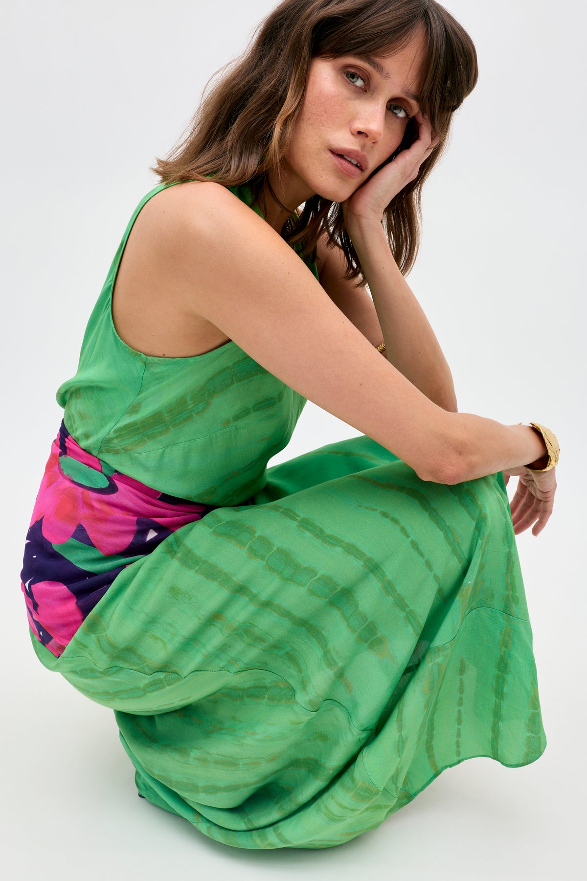Model wearing the Yousra - Tie Dye Green sleeveless dress with a colorful floral sash, seated on her heels and resting her head on her hand, gazing thoughtfully at the camera against a plain background.