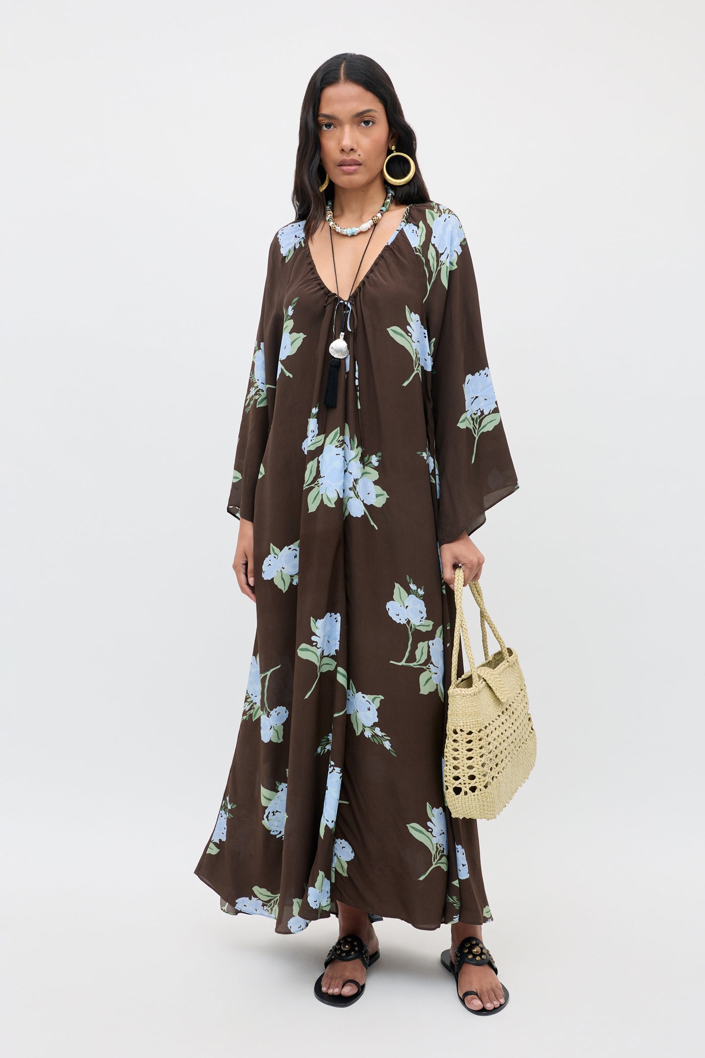 A woman stands against a plain white background wearing the "Lianne - Peonies Scatter Brown" long floral dress with blue flowers, paired with black sandals, layered necklaces, large hoop earrings, and a woven handbag.