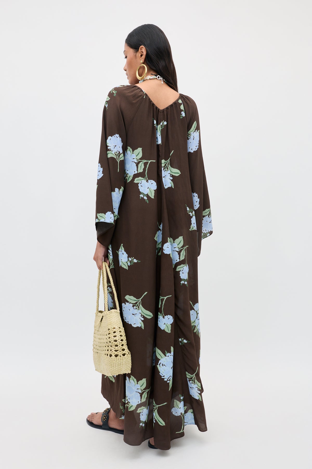 A woman stands facing away, wearing the Lianne - Peonies Scatter Brown dress—a long brown dress adorned with blue floral prints. She holds a woven straw bag, accessorizes with hoop earrings, and wears sandals.