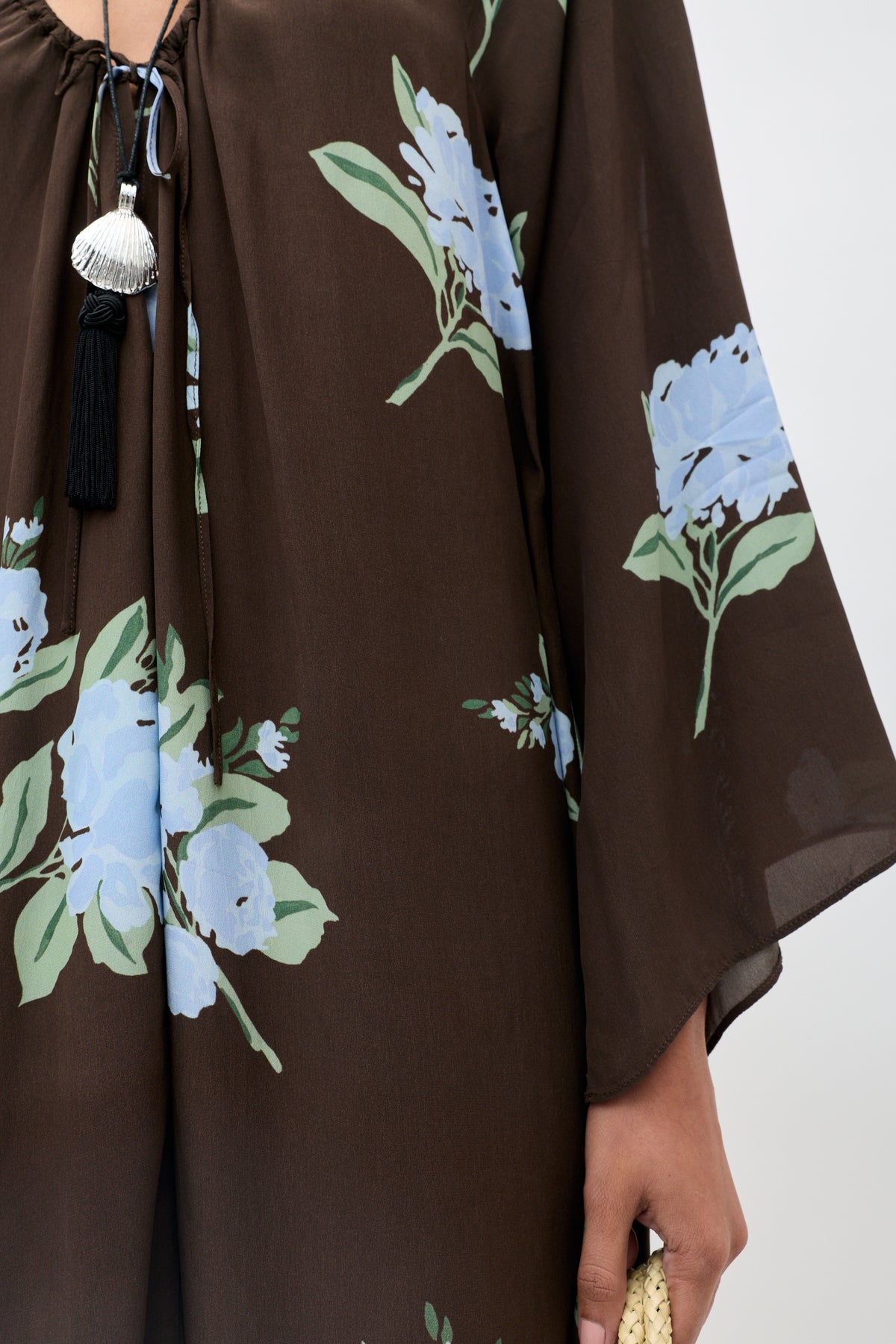 A person is shown holding a woven beige item while wearing the Lianne - Peonies Scatter Brown dress, which features a brown base with blue peony flowers and green leaves, adorned with black and silver tassels. Only their lower face, hand, and upper body are visible.
