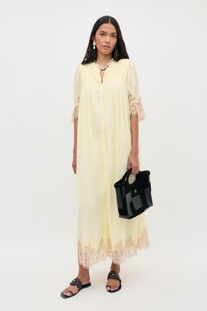 A woman with long dark hair stands against a plain white background wearing the Lorela - Lemon dress, a long, light yellow gown with lace trim and short sleeves. She accessorizes with a black handbag and black sandals.
