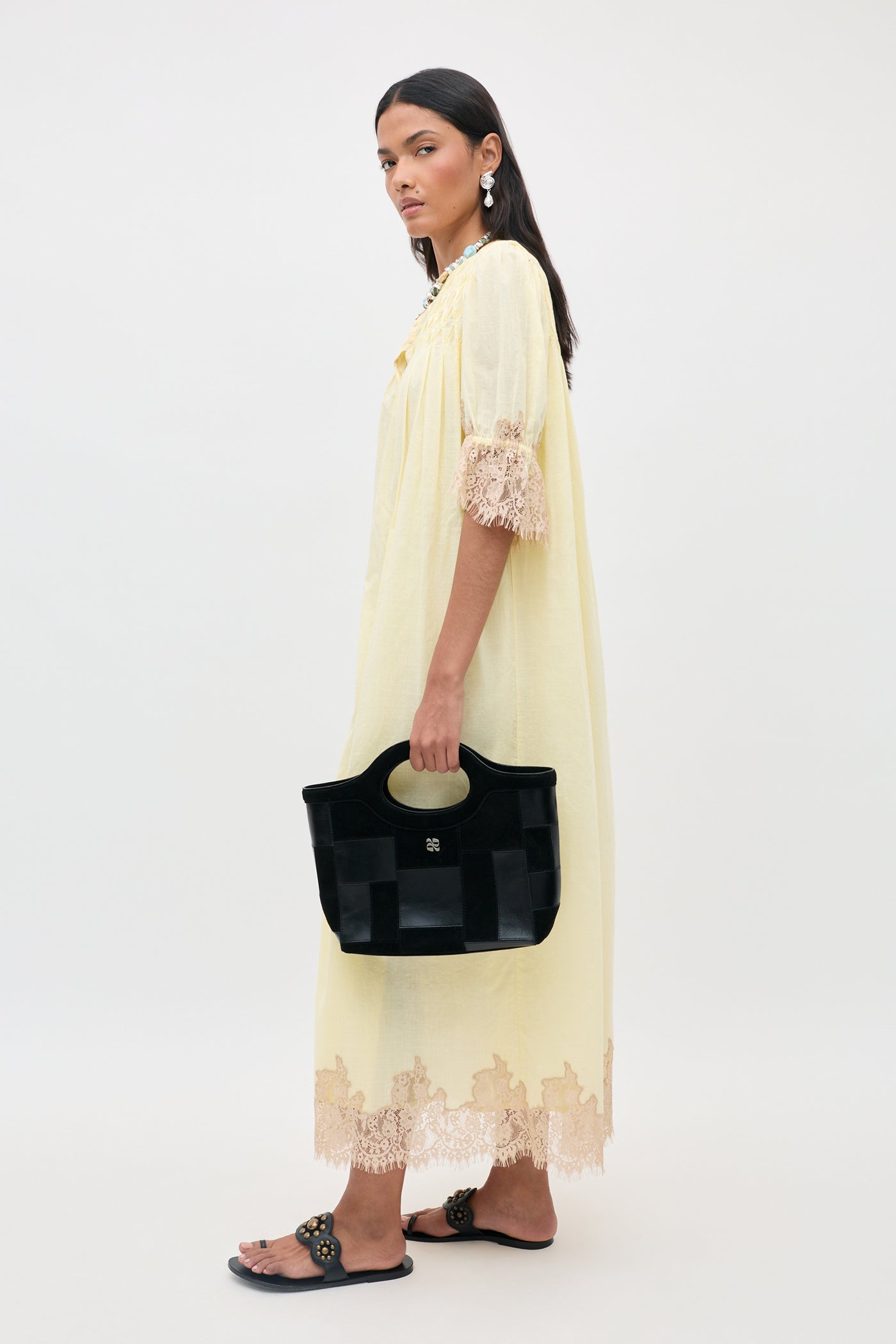 Woman in the Lorela - Lemon dress, featuring a light yellow hue and delicate lace trim, holds a black handbag. She pairs the look with embellished black sandals and statement earrings while standing against a plain white background.