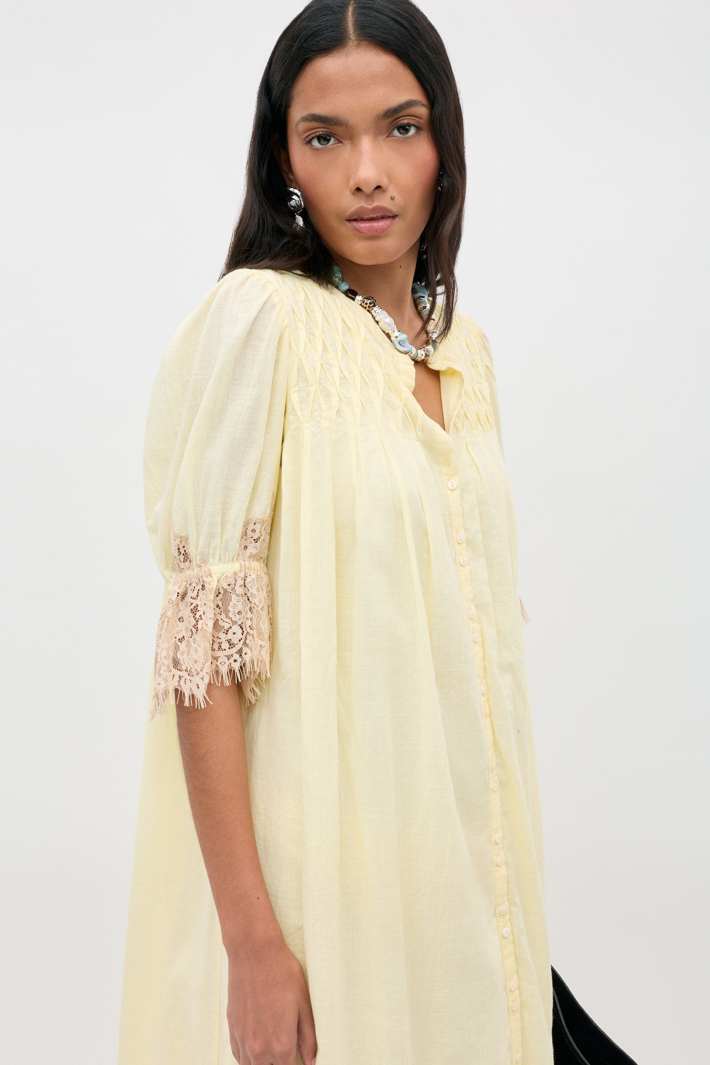 Wearing the Lorela - Lemon blouse—a pale yellow, loose-fitting top with lace-trimmed half sleeves and a button front closure—a woman accessorized with a silver necklace stands against a plain light background.