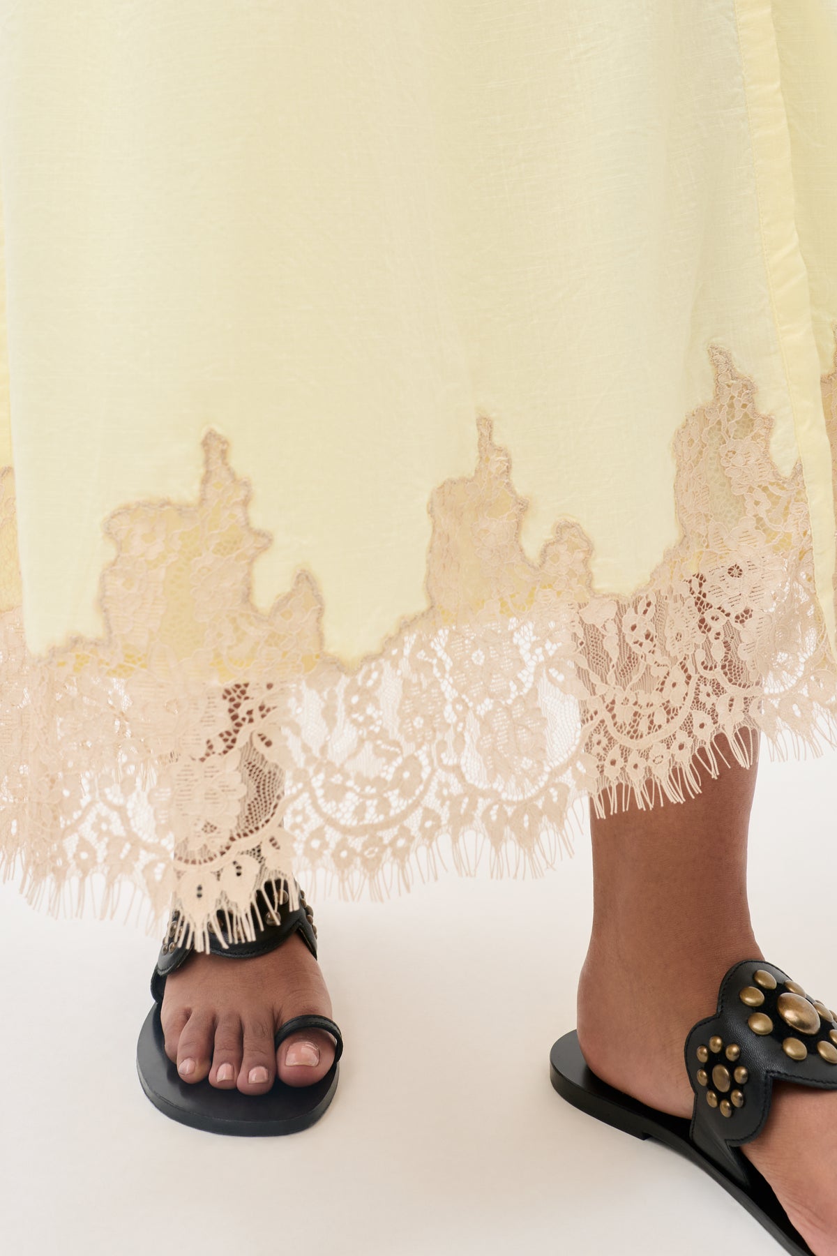 A close-up of a person wearing the Lorela - Lemon dress, a long, pale yellow piece featuring intricate lace trim at the hem, paired with black slide sandals adorned with gold studs.