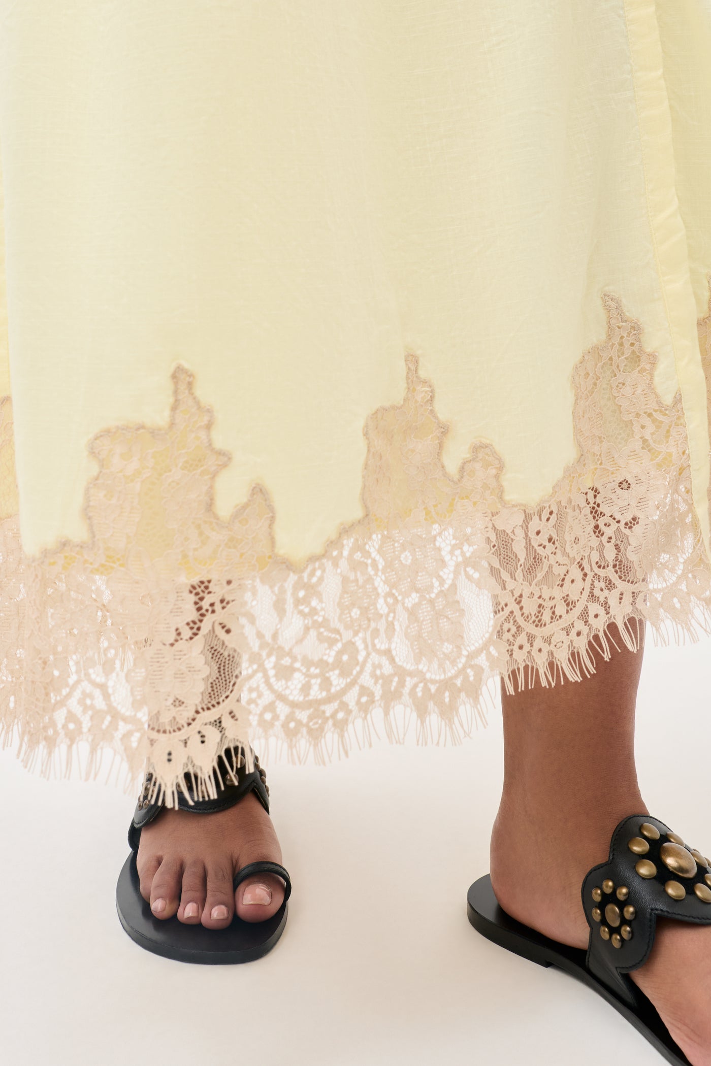A close-up of a person wearing the Lorela - Lemon dress, a long, pale yellow piece featuring intricate lace trim at the hem, paired with black slide sandals adorned with gold studs.