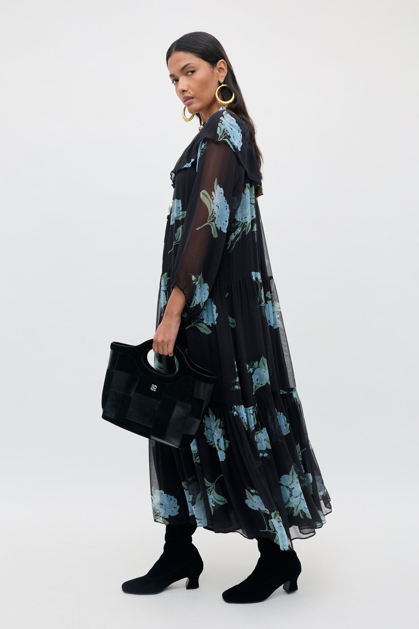 Woman in the Lela - Peonies Scatter Charcoal dress, featuring a sheer black fabric with blue floral patterns, styled with black ankle boots, gold hoop earrings, and a black handbag, standing against a plain light background.