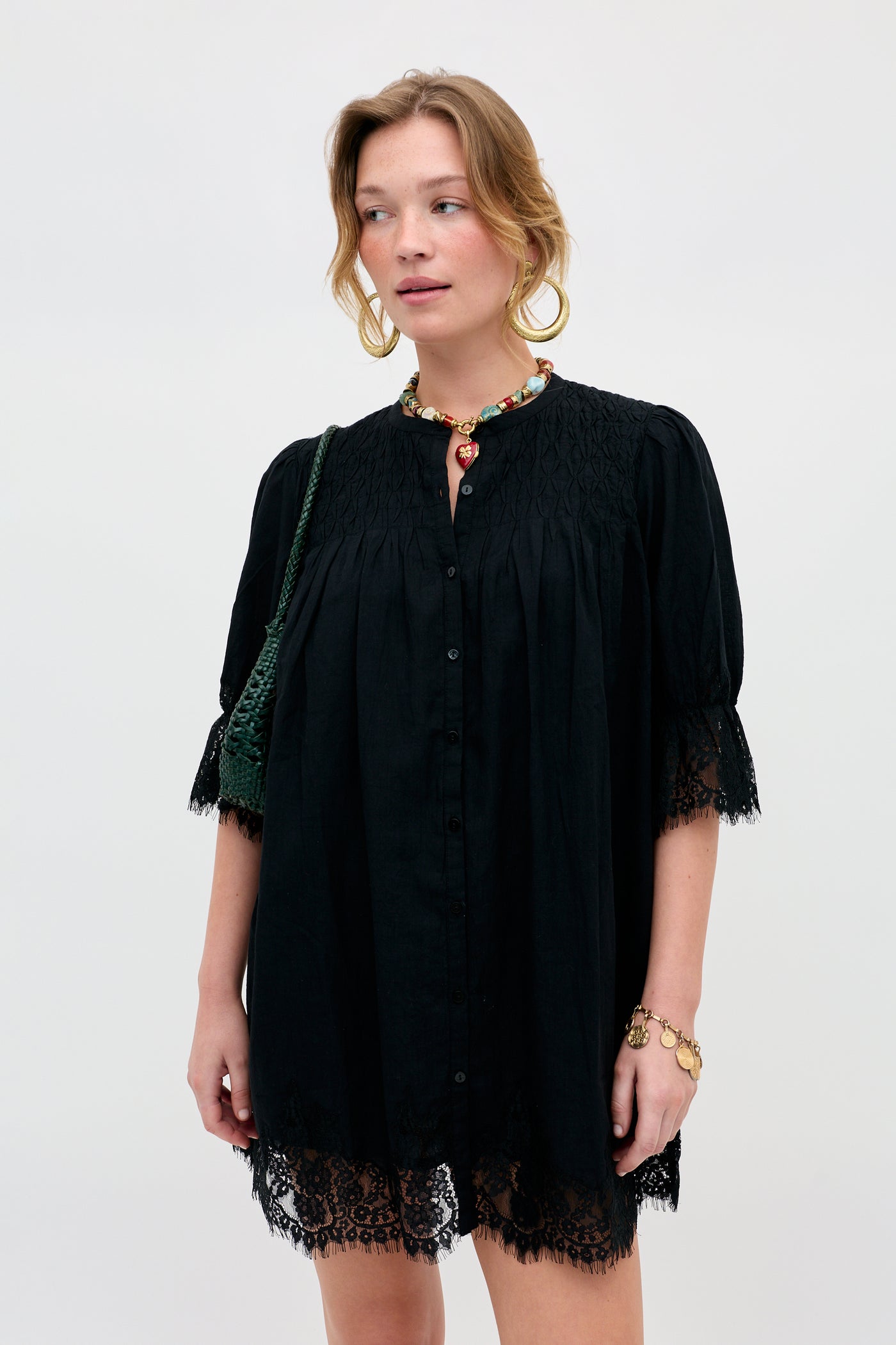 Woman wearing the Siera - Black loose black dress with lace trim, gold hoop earrings, a colorful necklace, and rings, holding a dark green bag, standing against a plain light background.