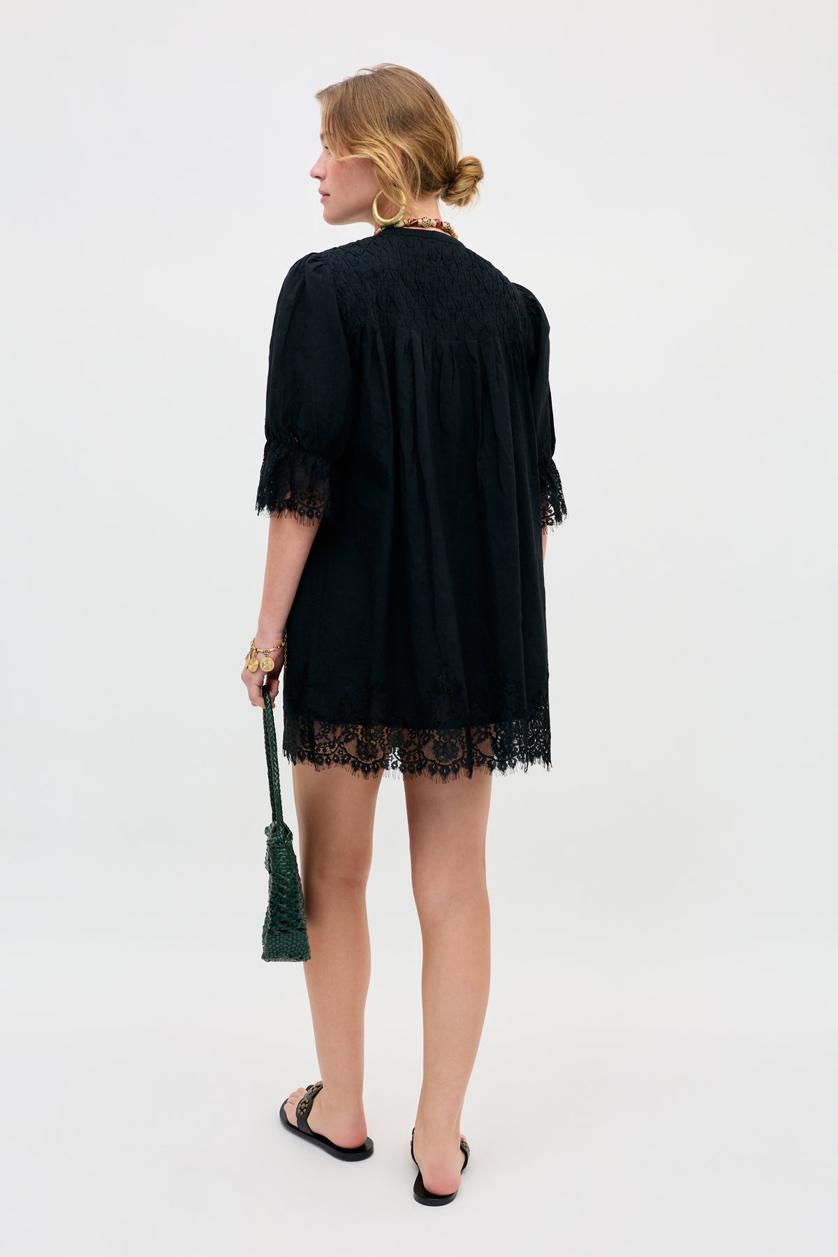 A woman stands facing away, wearing the Siera - Black dress with half sleeves. She holds a dark green tassel bag and wears flat black sandals. Her hair is styled in a low, loose bun as she poses against a plain, light background.