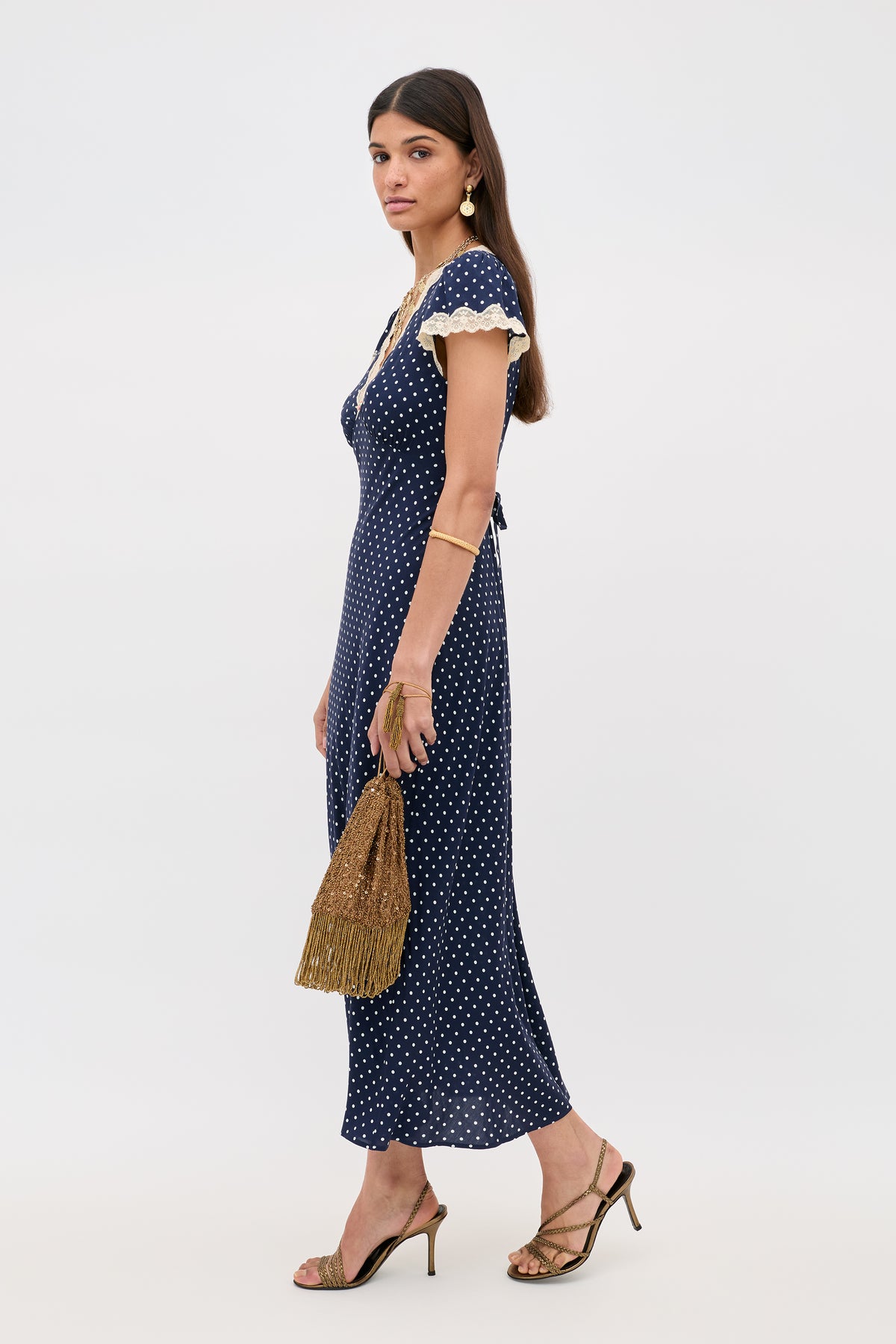 A woman models the Clarice - Navy Polka Dot rayon dress with lace trim, accessorized with a gold fringed handbag, gold earrings, bracelets, and heeled sandals against a plain white background.