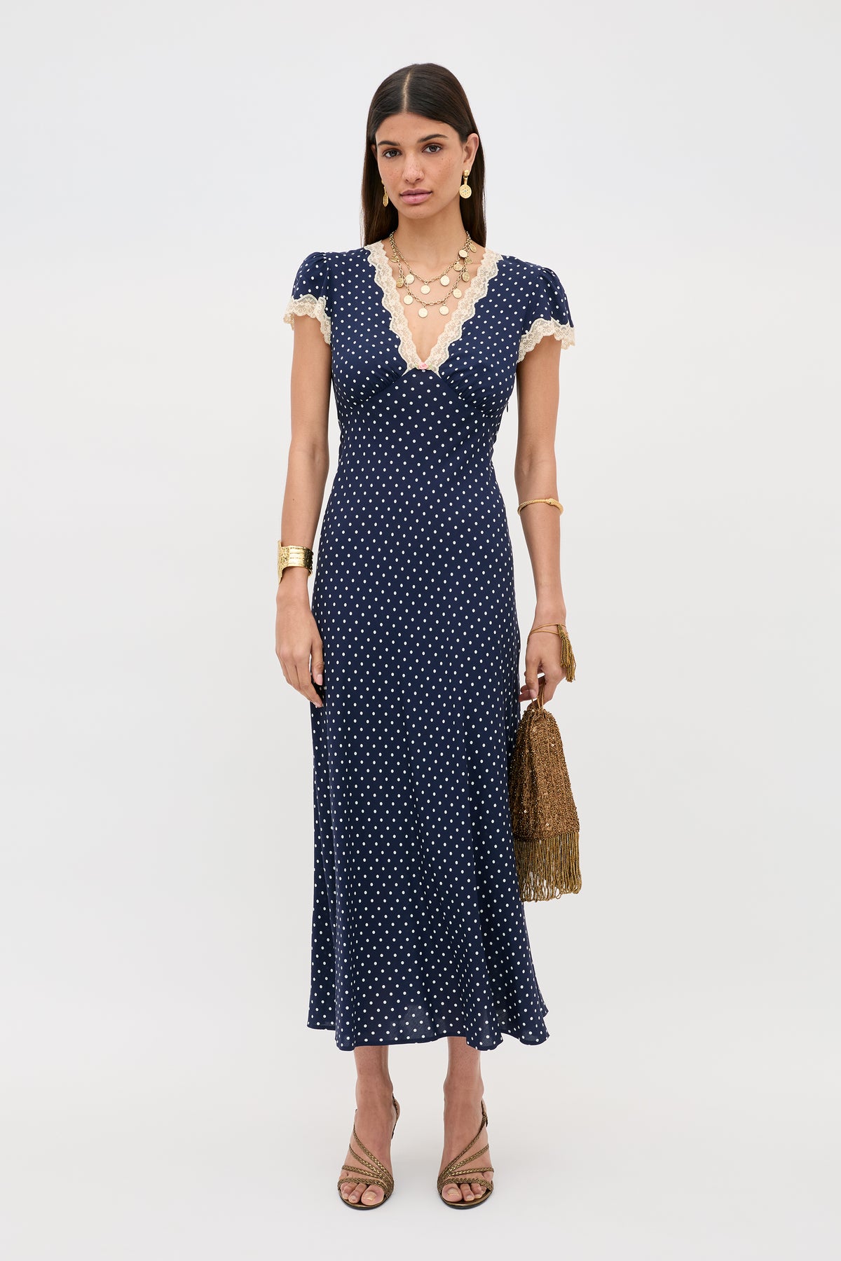 A woman stands against a plain background wearing the Clarice - Navy Polka Dot dress—a rayon, deep V-neck style with white lace trim—paired with tan heels, layered necklaces, gold bracelets, and a brown fringe handbag.