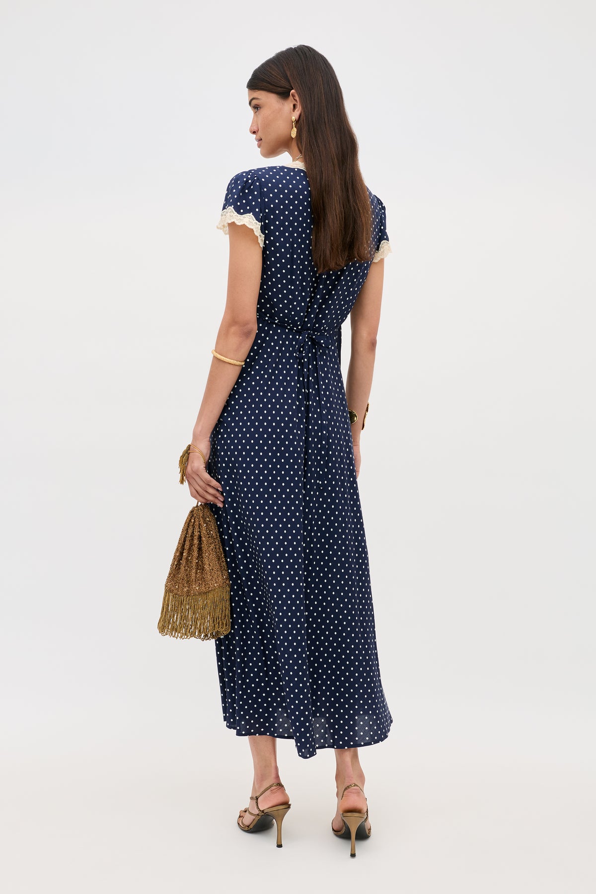 A woman with long dark hair faces away, wearing the Clarice - Navy Polka Dot ankle-length rayon dress with lace trim. She holds a gold fringe handbag and accessorizes with heeled sandals and gold jewelry.