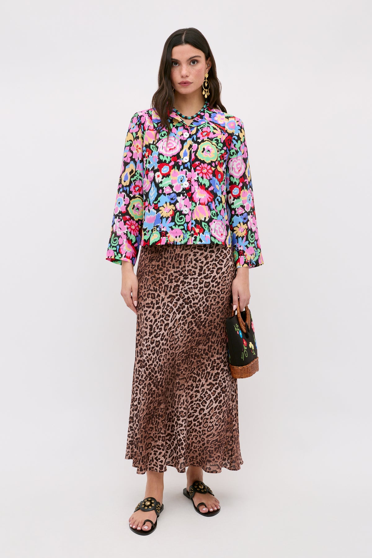 A woman stands wearing a colorful floral blouse, the Kelly - Leopard Brown skirt crafted from luxurious silk crepe de chine featuring a long leopard print design, black sandals, and holding a small handbag. She has long dark hair and wears gold earrings against a plain light background.