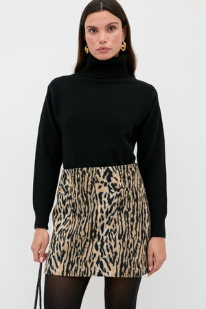 A woman in a black turtleneck, black tights, gold earrings, and the Mariah - Bohemia Leopard Brown mini skirt stands against a white background, holding a black purse—serving up chic noughties style.