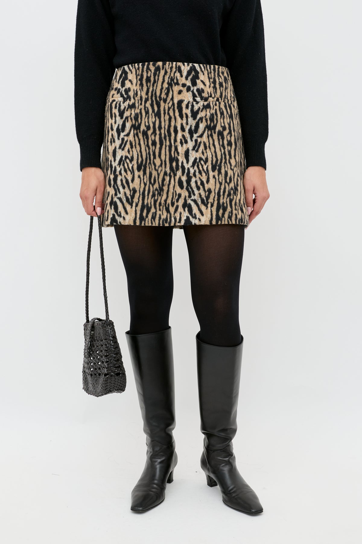 Wearing the Mariah - Bohemia Leopard Brown mini skirt, black sweater, black tights, knee-high boots, and holding a textured black handbag, a person stands against a plain white background.