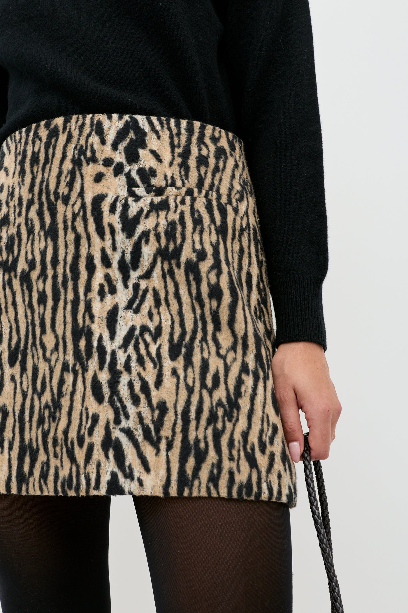 A person wearing a black long-sleeve top, black tights, and the Mariah - Bohemia Leopard Brown mini skirt, holding a black textured strap in their right hand.