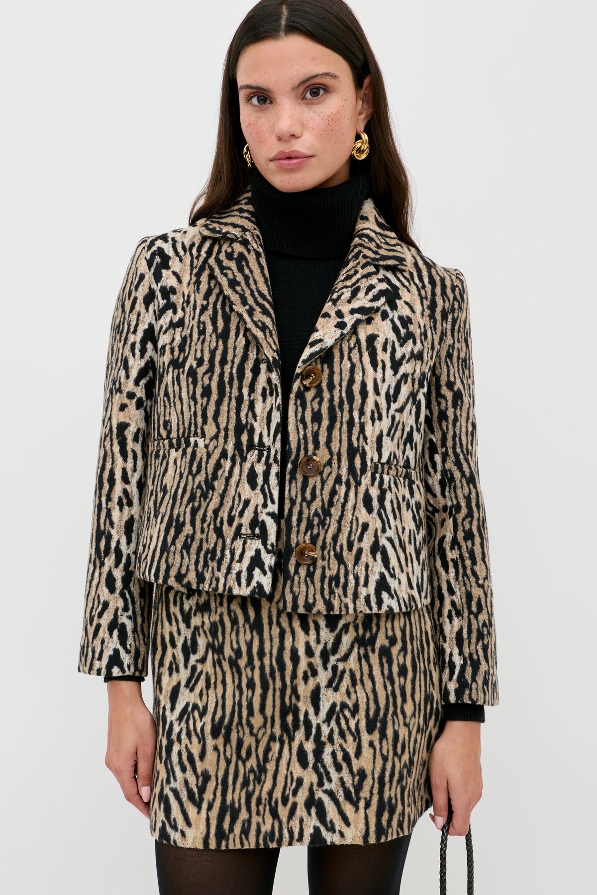 Wearing the Mariah - Bohemia Leopard Brown set, a woman pairs a black turtleneck with the leopard-print mini skirt and jacket, accessorizing with gold earrings and a small bag for a bold noughties-inspired look against a white background.