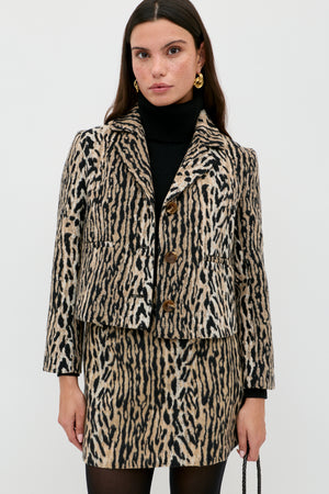 Wearing the Mariah - Bohemia Leopard Brown set, a woman pairs a black turtleneck with the leopard-print mini skirt and jacket, accessorizing with gold earrings and a small bag for a bold noughties-inspired look against a white background.