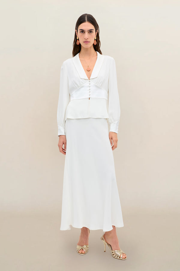 A woman wears Joan - Ivory, a long-sleeved white blouse with front buttons paired with a matching satin-back silk maxi skirt. She accessorizes with gold earrings, a necklace, and gold open-toe heels. The background is plain light beige.