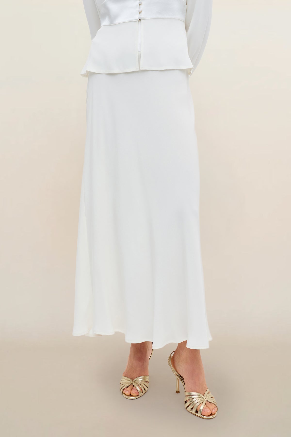 A person wearing the Joan - Ivory satin-back silk maxi skirt and gold strappy high-heeled sandals, standing against a neutral background.