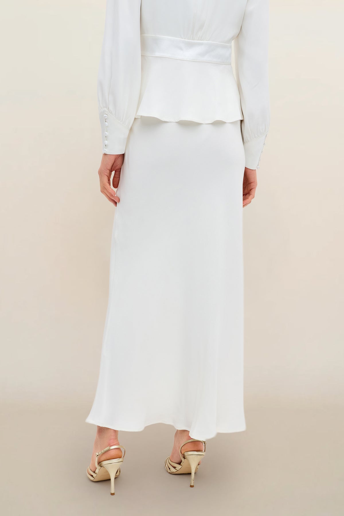 A person is shown from the back standing on a neutral background, wearing Joan - Ivory—a long ivory satin-back silk maxi skirt with a matching long-sleeve top—and gold high-heeled sandals.