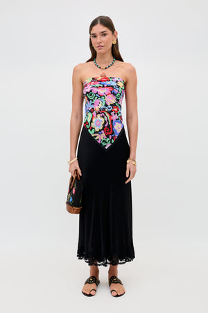 A woman wears the Crystal - Black dress, which features a colorful floral bodice and a flowing black satin midi skirt with delicate lace trim. She accessorizes with sandals, a necklace, a bracelet, and a handbag while standing against a plain white background.