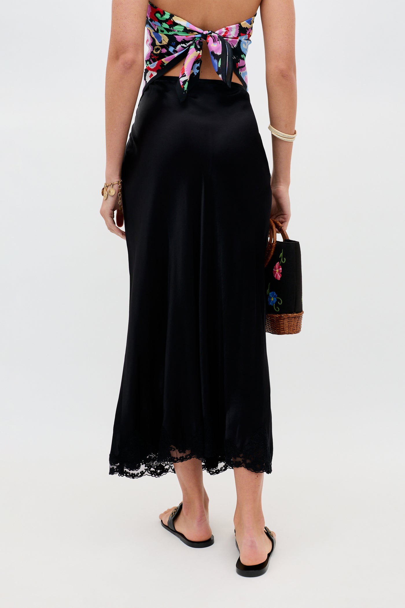 A woman stands facing away, wearing the Crystal - Black satin midi skirt with delicate lace trim cut on the bias, paired with a colorful back-tie top, black sandals, and holding a black handbag with floral embroidery and a wicker base.