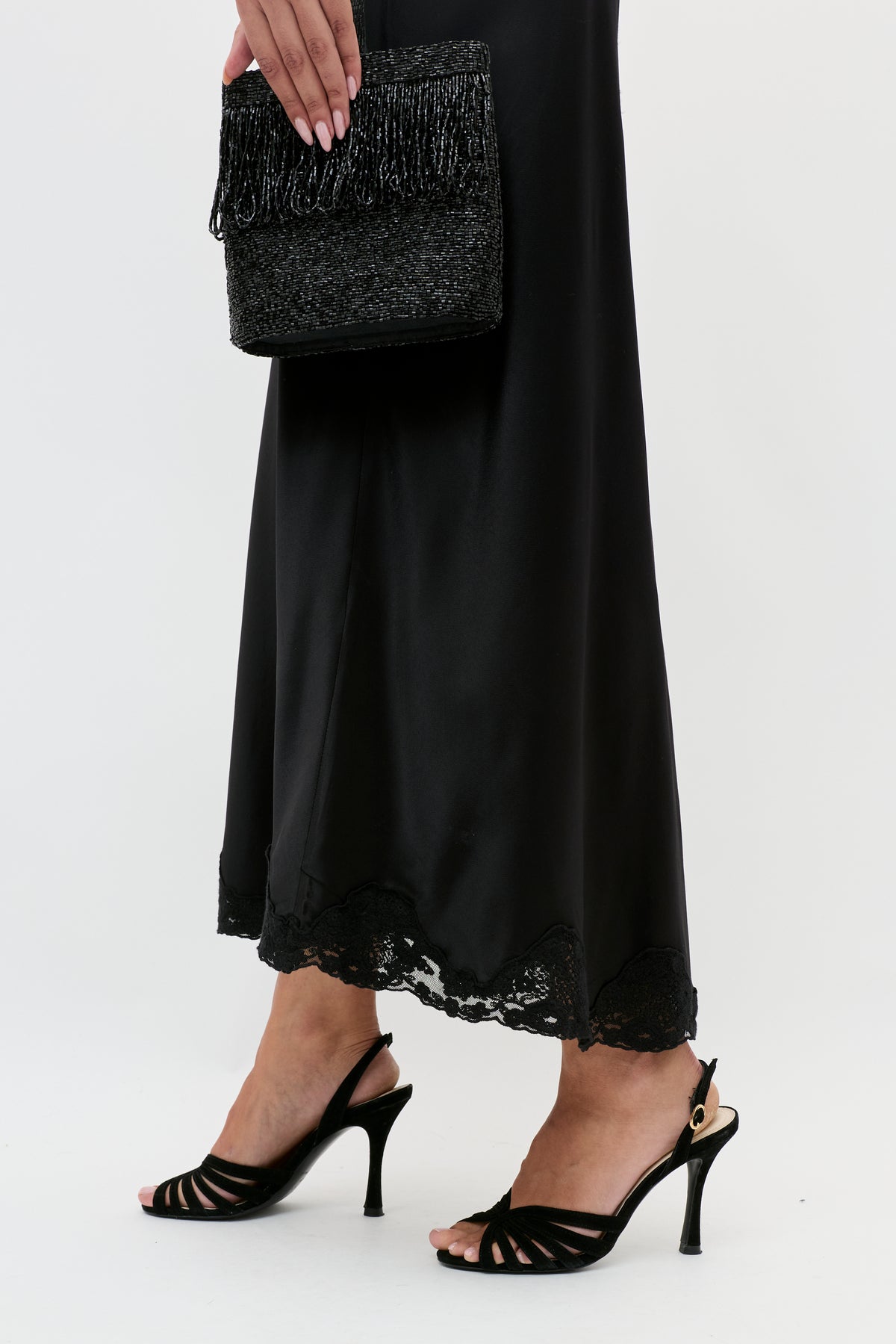 Wearing the Crystal - Black satin midi skirt with lace trim and black heels, a person holds a textured black clutch with fringe, standing against a white background.