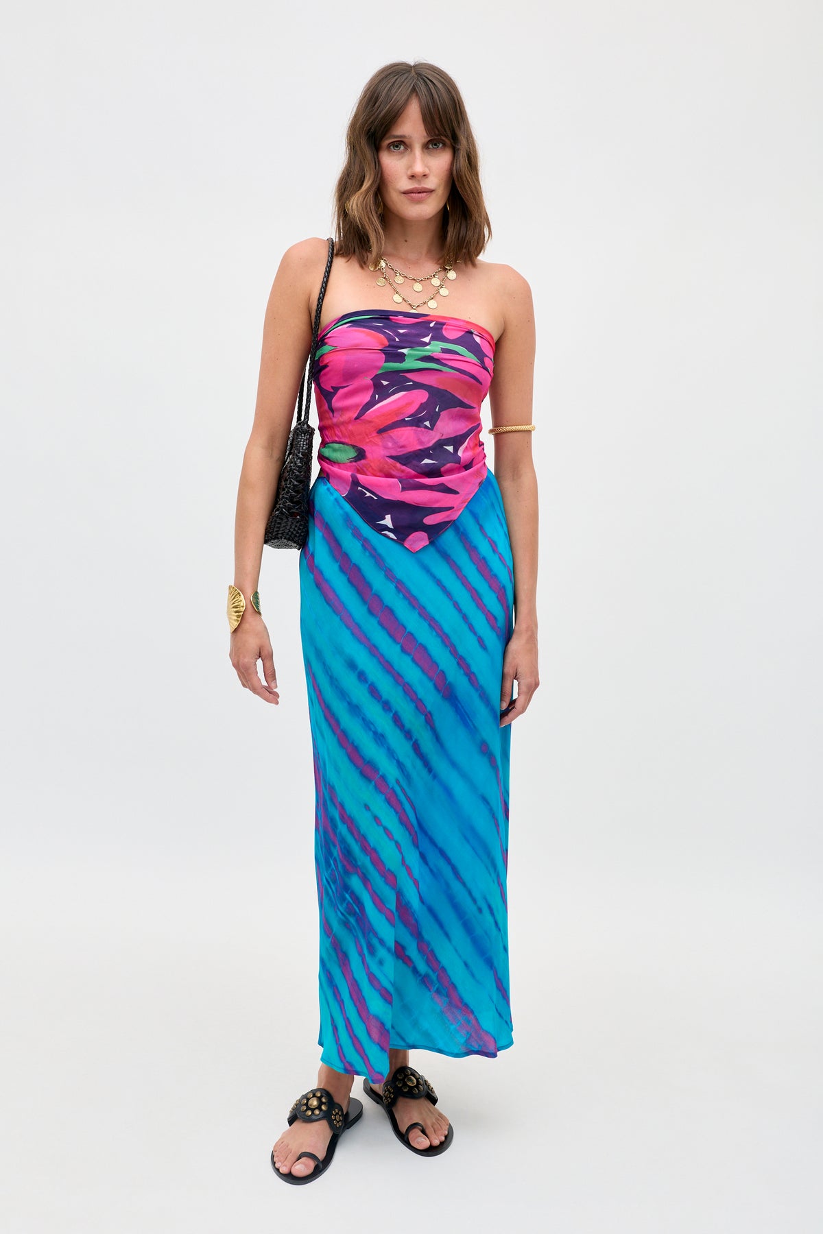 A woman wearing the Ardith - Tie Dye Blue dress, a strapless piece featuring a vibrant pink floral print on the top and blue diagonal stripes on the skirt, styled with layered necklaces, bracelets, a black purse, and black sandals.
