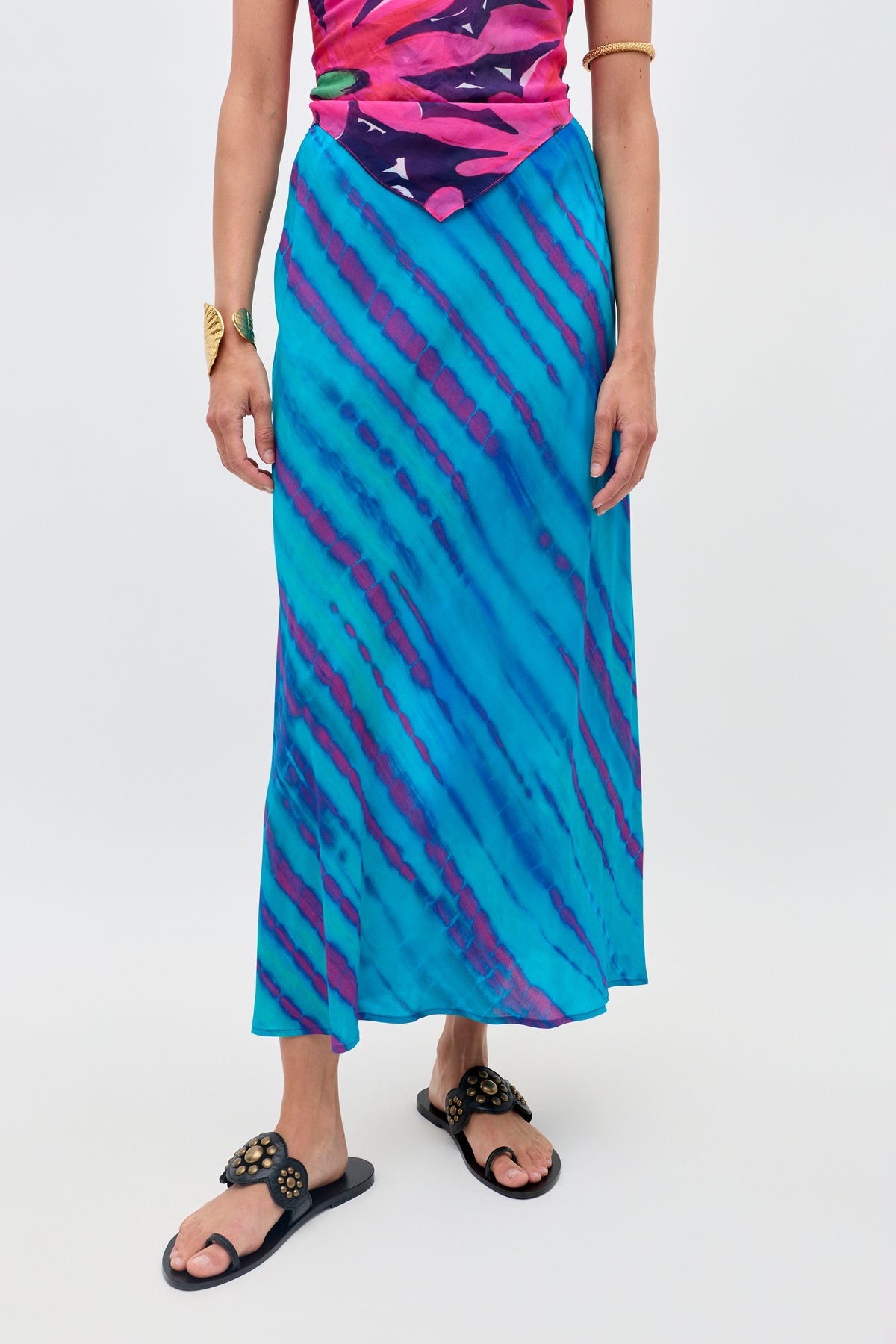 A person wearing the Ardith - Tie Dye Blue long skirt with purple diagonal stripes, paired with a colorful pink and purple top, black sandals, and gold bracelets stands against a plain background.