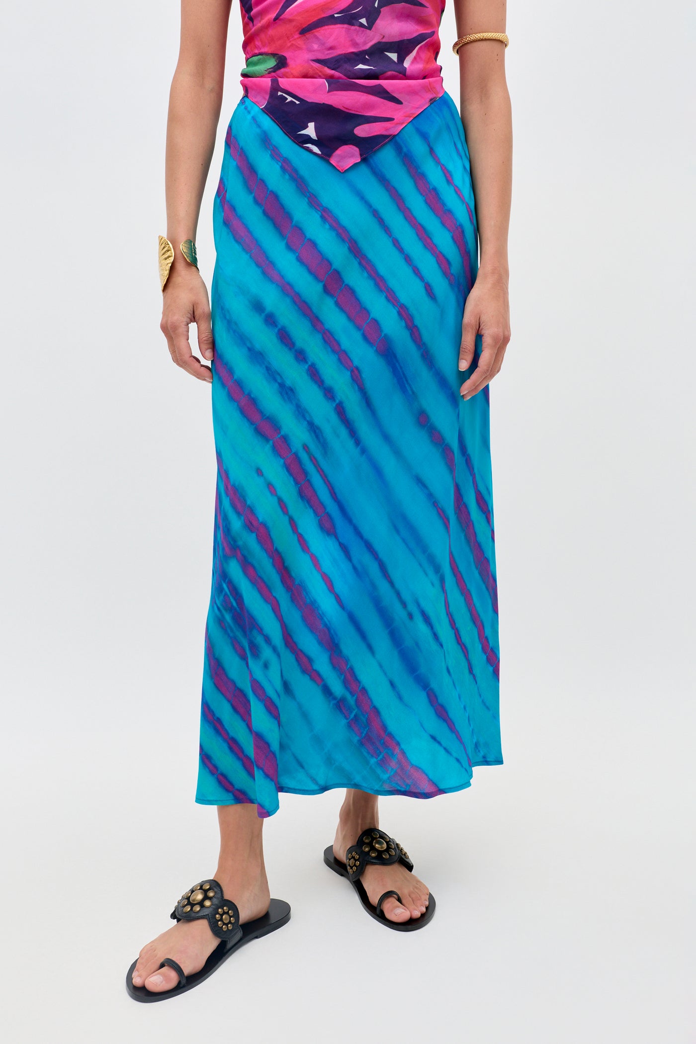 A person wearing the Ardith - Tie Dye Blue long skirt with purple diagonal stripes, paired with a colorful pink and purple top, black sandals, and gold bracelets stands against a plain background.