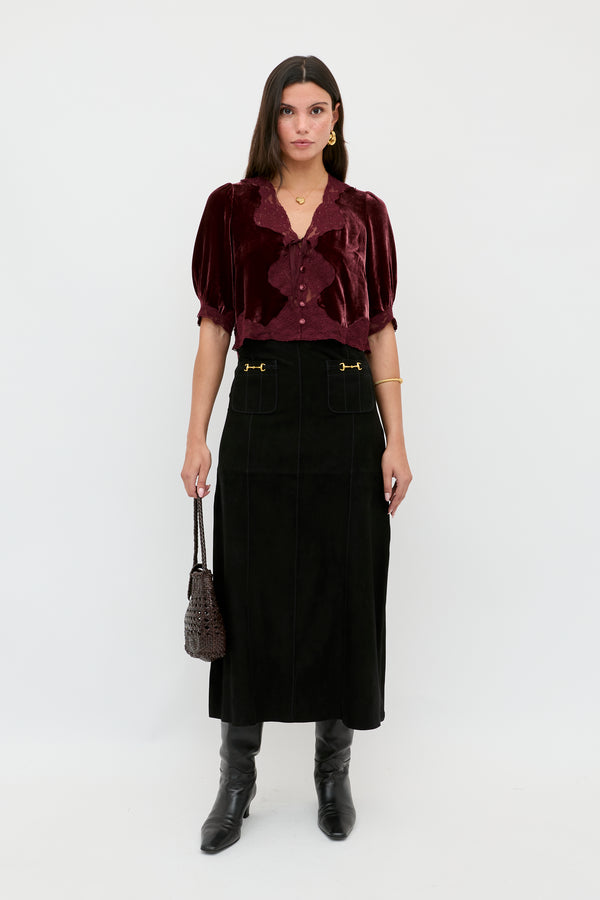A woman with straight brown hair and a serious expression stands against a white background wearing the Edwina - Black high-waisted midi skirt with gold accents, a burgundy velvet blouse, black knee-high boots, and holding a small dark handbag.