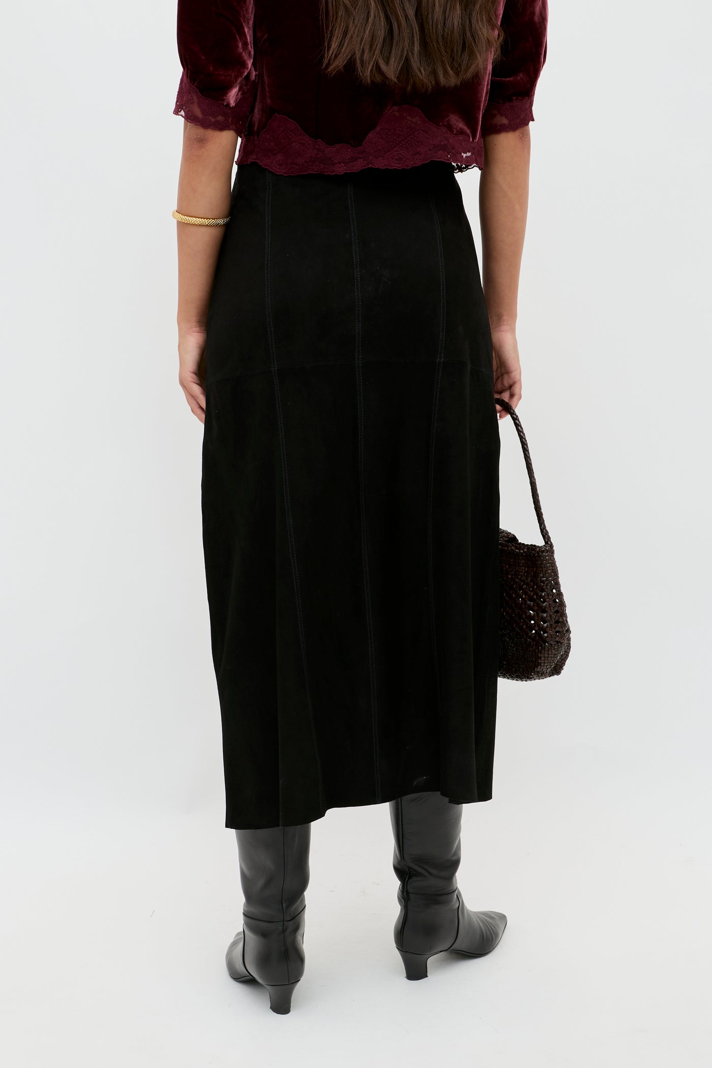 A person stands facing away, wearing the Edwina - Black high-waisted A-line skirt, paired with a burgundy velvet top, black knee-high boots, and holding a brown woven bag against a plain light background.