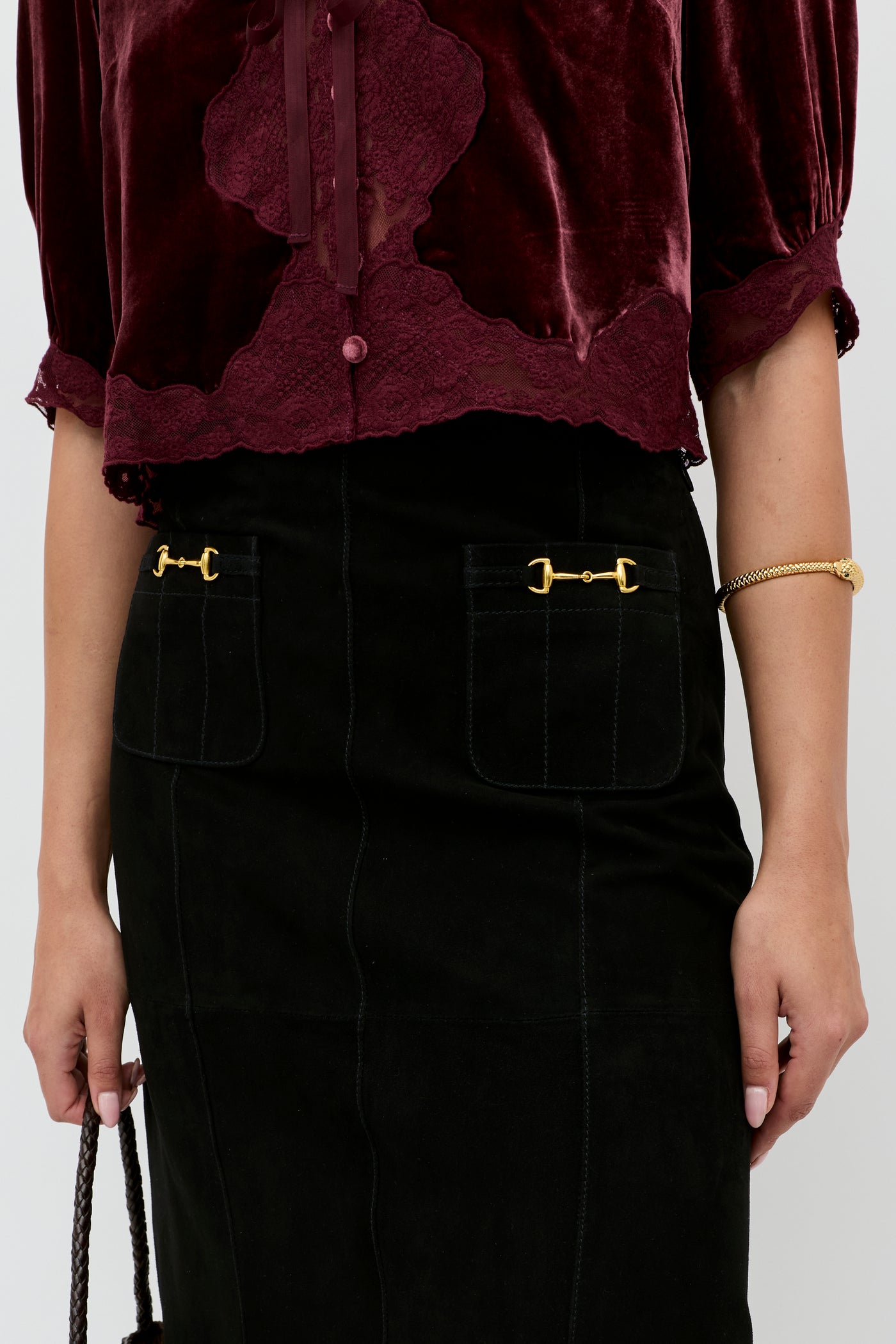 A person is wearing a burgundy velvet blouse with lace trim, the Edwina - Black high-waisted skirt featuring gold decorative hardware on the pockets, and a gold bracelet. Only their torso and arms are visible.