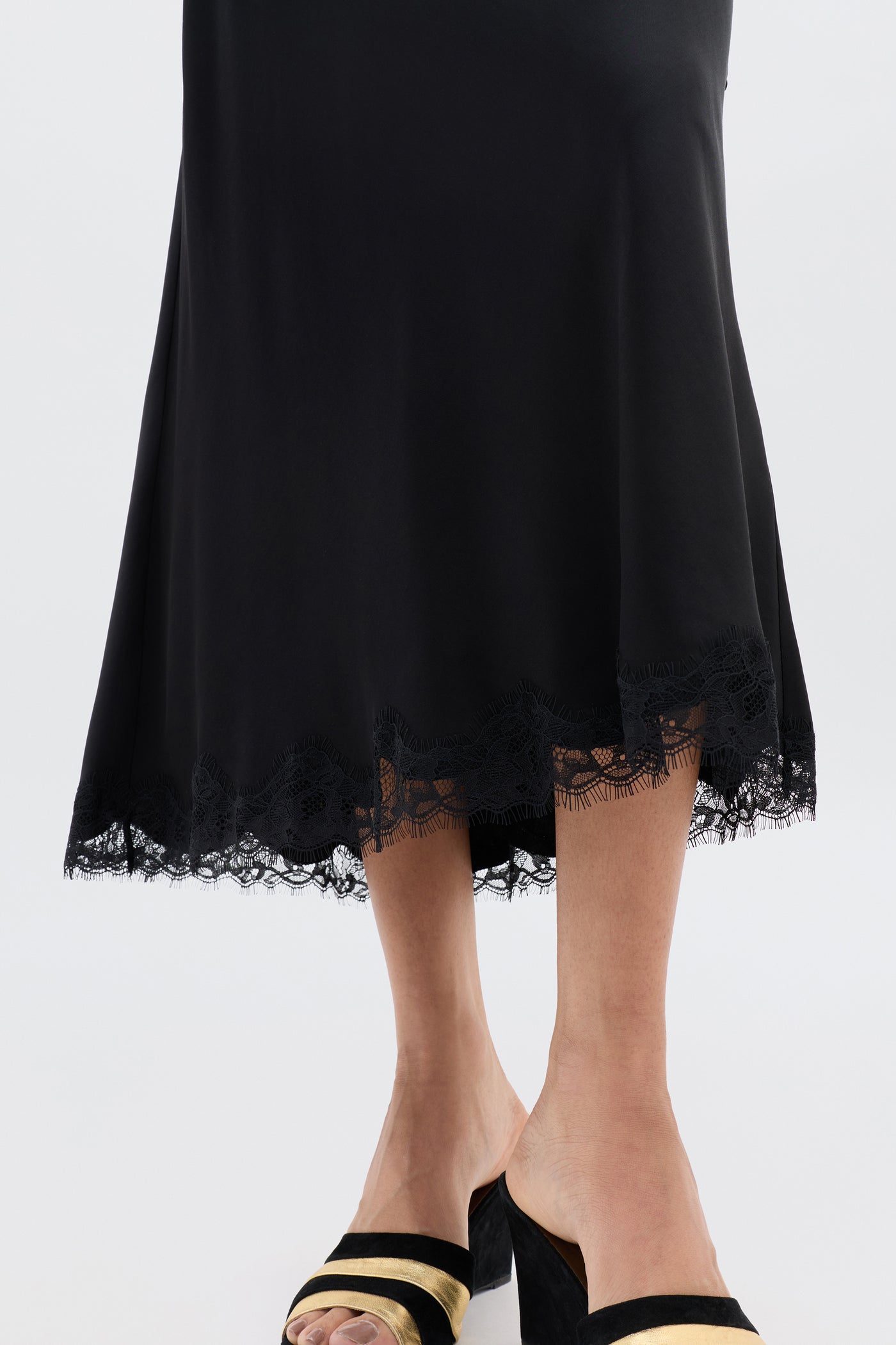 A person wearing the Stassie - Black satin skirt with wavy lace trim at the hem and black open-toe heels with gold accents, standing against a plain light background.
