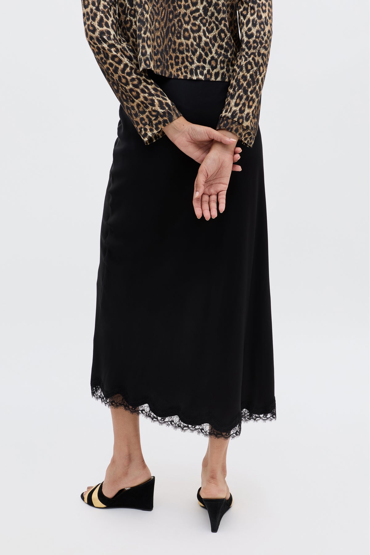 Clad in the Stassie - Black skirt with wavy lace trim, a leopard print long-sleeve top, and black wedge sandals, the person stands hands clasped behind their back, channeling effortless Parisian style against a plain backdrop.