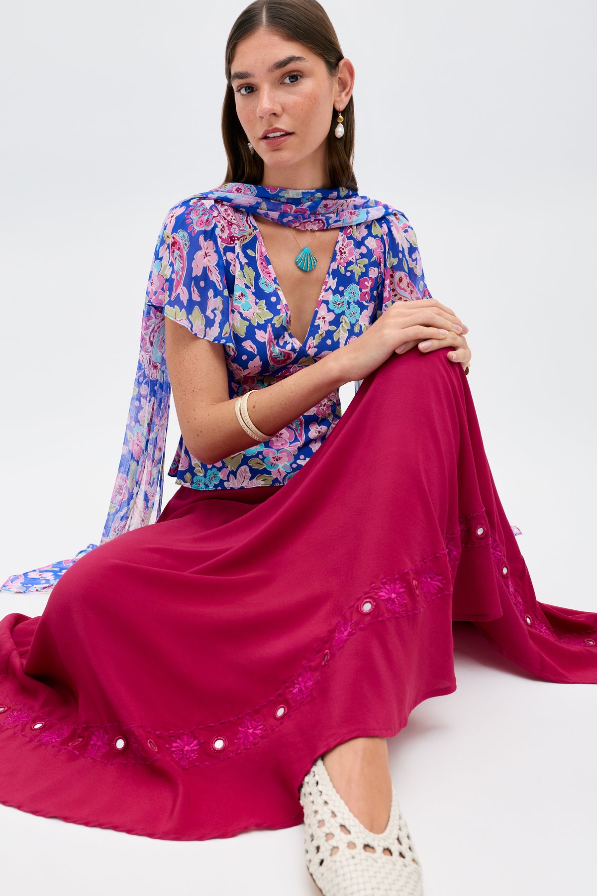 A woman sits on the floor wearing a floral top with a matching scarf, accessorized with earrings and a turquoise necklace. She pairs her outfit with the ‘Rudy - Mirror Embellishment Magenta Pink’ skirt featuring lace details and white woven shoes, her arms resting on her knee.